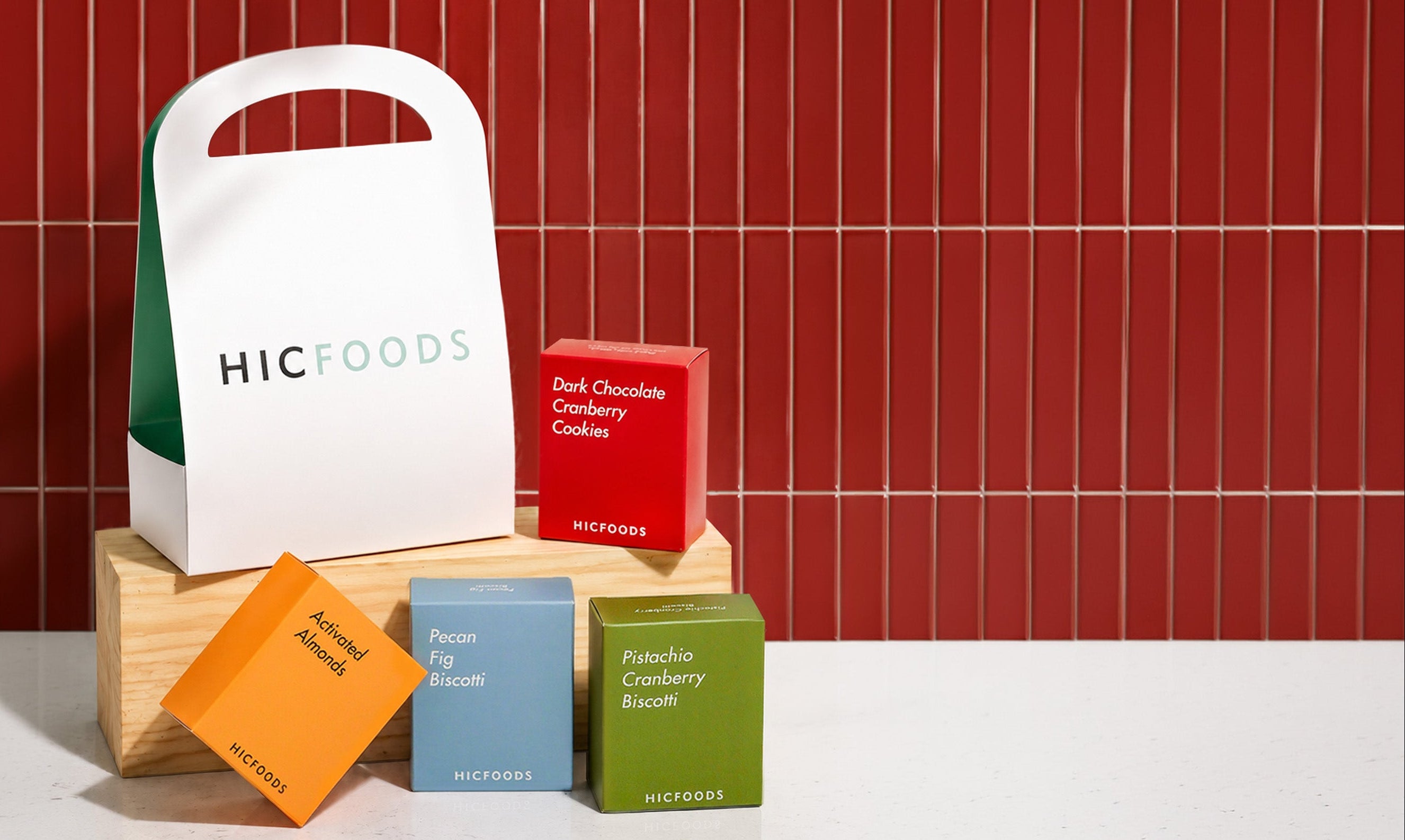 A white HICFOODS paper gift bag with a handle stands on a wooden block beside several colorful product boxes, including red, orange, blue, and green boxes, against a red tiled wall and white countertop.